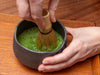 Person whisking matcha green tea in a black bowl on a wooden surface. Usucha.