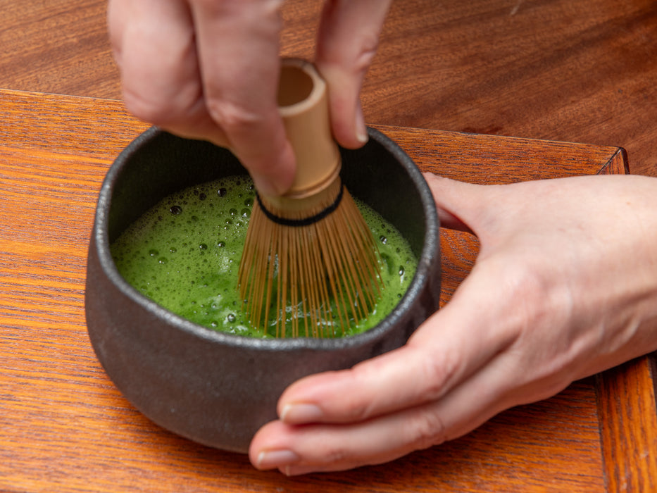 Person whisking matcha green tea in a black bowl on a wooden surface. Usucha.